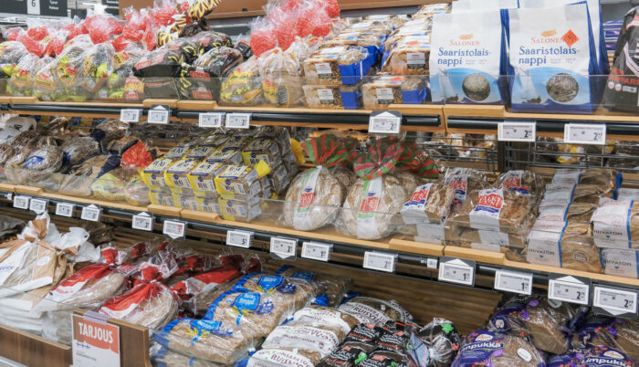 A row of grocery store shelves contains dozens of different kinds of dark bread in plastic bags.