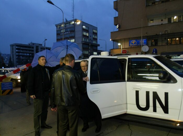 Several people holding umbrellas wait as a man steps out of an automobile marked with the abbreviation for United Nations.