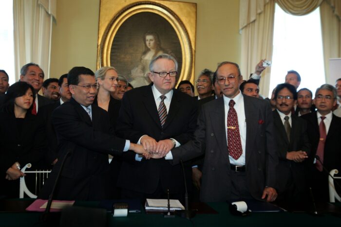 In a stately room, two men are shaking hands, while another man has laid his hands on top of their hands in agreement. In the background is a crowd of dozens of people.