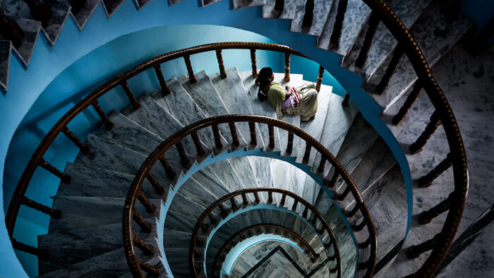 Plusieurs étages desservis par un escalier en colimaçon orné sont visibles depuis un étage supérieur, tandis qu’une femme est assise sur l'une des marches de l’escalier.