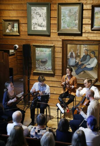 在一间墙壁上挂着许多画作的房间里，一名女性在唱歌，另一名女性和三个男性在为观众演奏弦乐器。