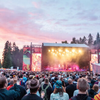 With the sky showing pink and orange sunset colours, thousands of festivalgoers watch a concert on a large outdoor stage flanked by a forest.