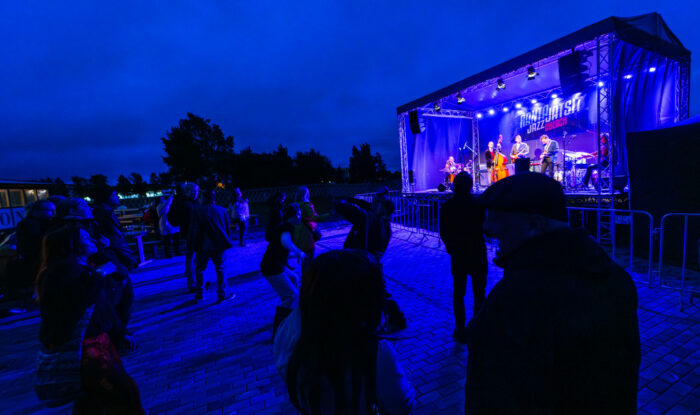 Tandis que la nuit tombe, des gens assistent à un concert en dansant devant une scène en plein air.