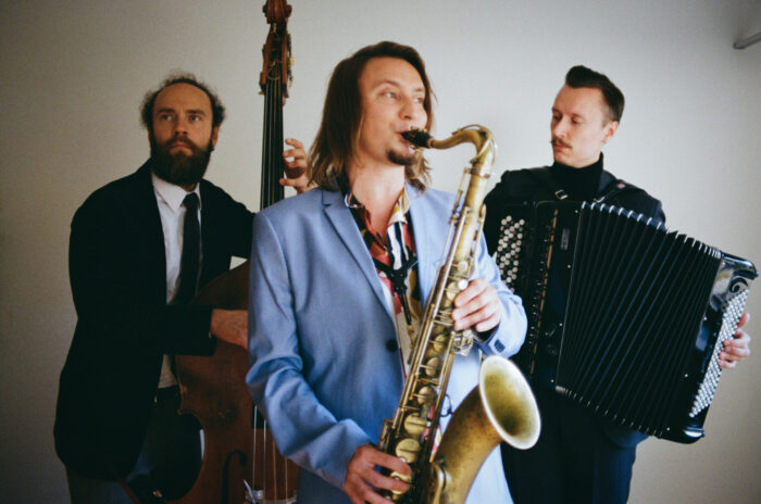 Three men are standing in a row, playing double bass, tenor saxophone and accordion.