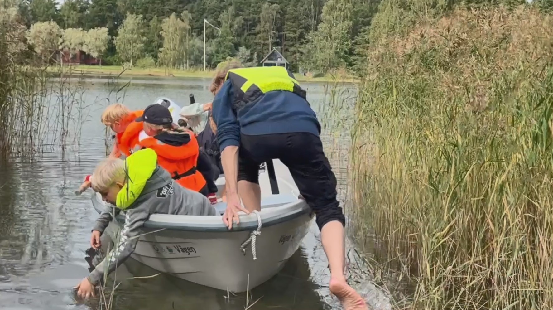 A man is hopping into a boat with several children in it while he pushes it away from the shore with one foot.