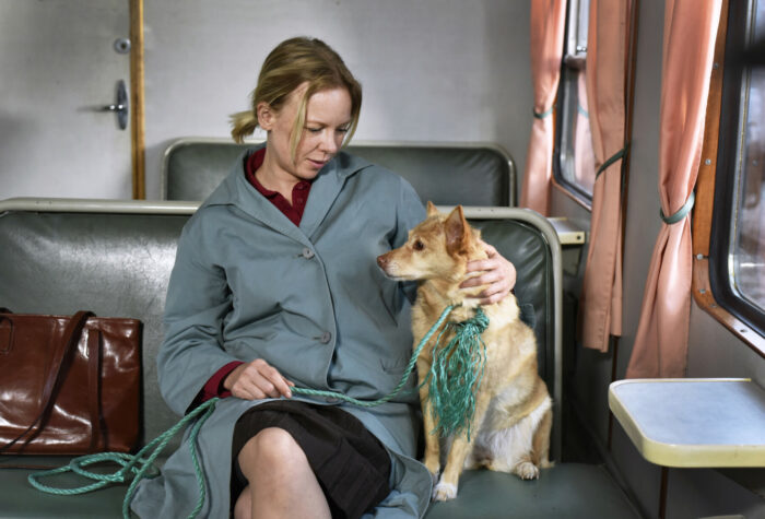 In a commuter train, a woman and a dog sit on a bench, the woman with her arm around the dog.