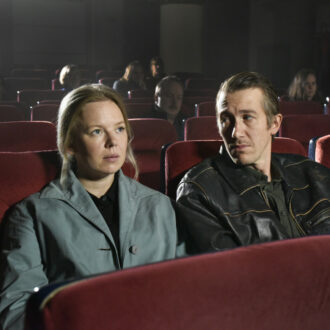 Uma mulher e um homem estão sentados num cinema, o homem vira a cabeça para olhar para a mulher que está assistindo ao filme.
