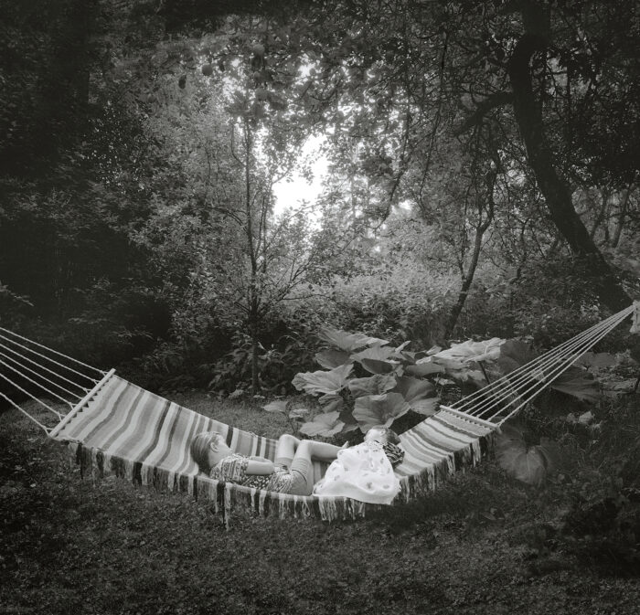 Two children sleep in a hammock under leafy trees.