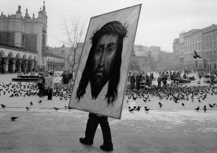 A huge portrait of Jesus's head is being carried across a town square by someone who is hidden behind the portrait except for a pair of legs that are visible.