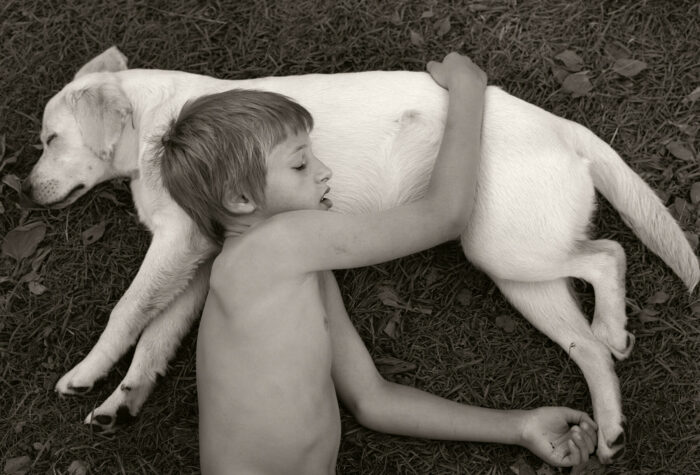 Um menino dorme com a cabeça e o braço apoiados em um cachorro que também está dormindo.