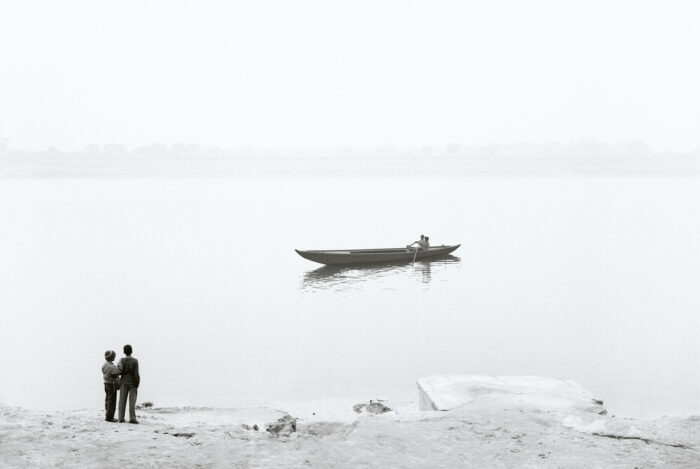 En la orilla de un lago, dos personas observan el paso de una embarcación parecida a una canoa.