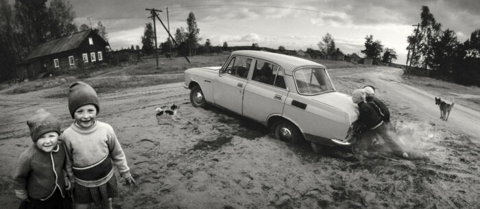 Plusieurs jeunes garçons poussent une voiture embourbée dans de la terre boueuse, tandis qu'au premier plan deux petites filles sourient à l’objectif du photographe.