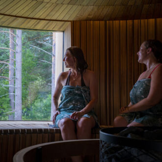 Two women wrapped in towels sit on a bench in a sauna, looking out the window at a forest.