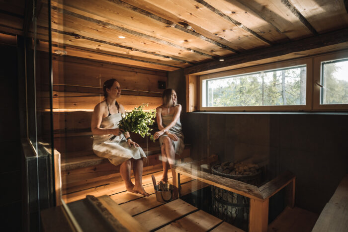Two women wrapped in towels sit on a bench in a sauna, looking out the window at a forest.