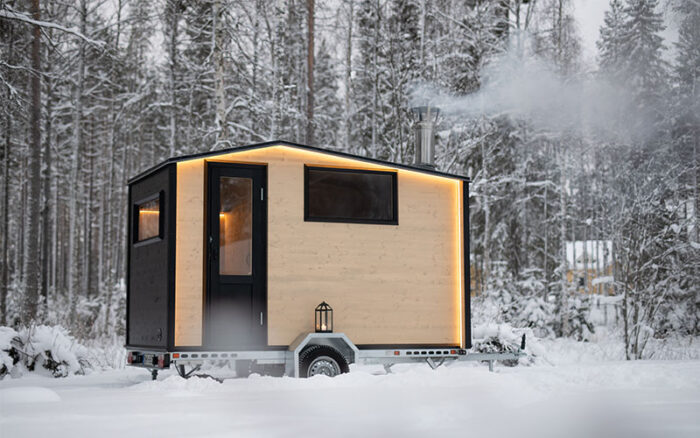 A small wooden cabin on a trailer bed with wheels, parked in a snowy forest landscape.