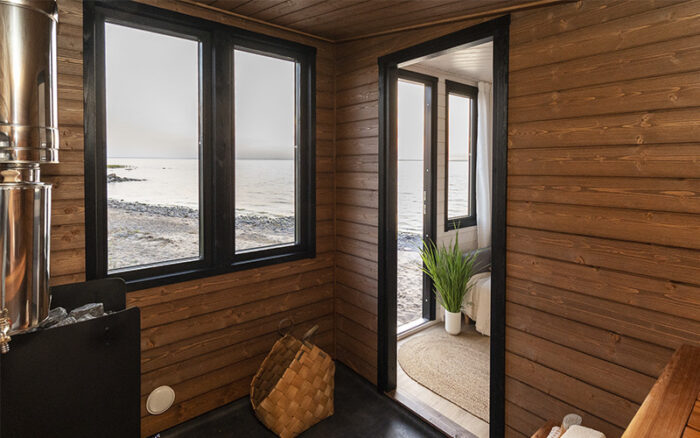 On voit l'intérieur d'un sauna avec une porte ouverte sur un vestiaire et une fenêtre donnant sur une plage.