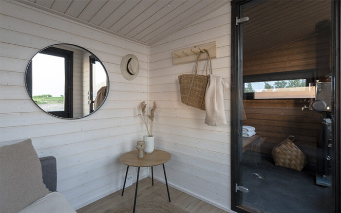 On voit un vestiaire avec une porte vitrée donnant sur un sauna et un miroir où se reflète un paysage baigné par un plan d’eau.