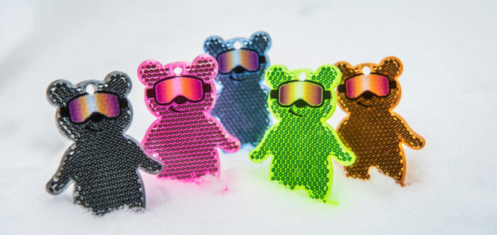 Five small bear-shaped pieces of plastic of various colours are standing in a pile of snow.