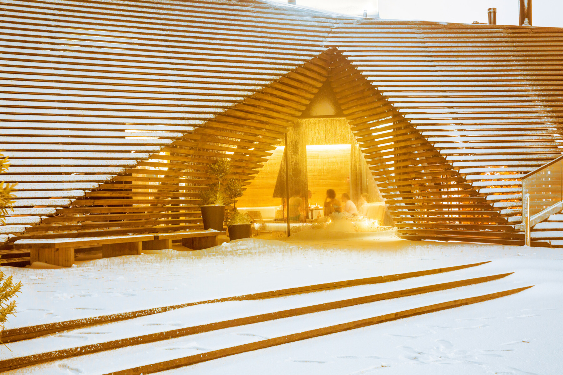 A wooden building with a triangular archway is snowy on the outside and a window shows people sitting in a cosy room on the inside.