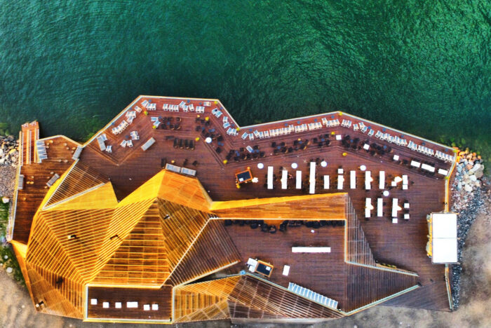 An aerial shot shows a modern, angular wooden building with several patios beside the sea.