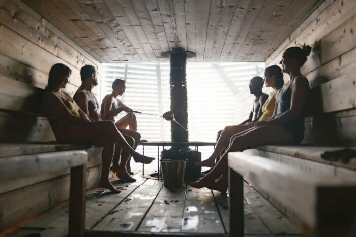 Шесть человек сидят на скамьях сауны, через окно в стене в сауну проникает естественный свет.