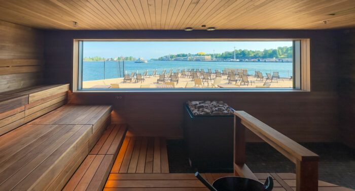 On voit un sauna doté de plusieurs bancs en bois disposés en gradins et d’une large baie vitrée rectangulaire offrant une vue sur la mer.