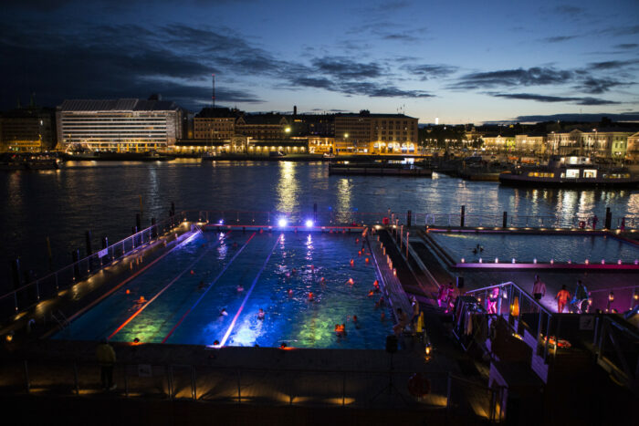 On one side of a harbour at twilight, rows of lights show where several swimming pools are.