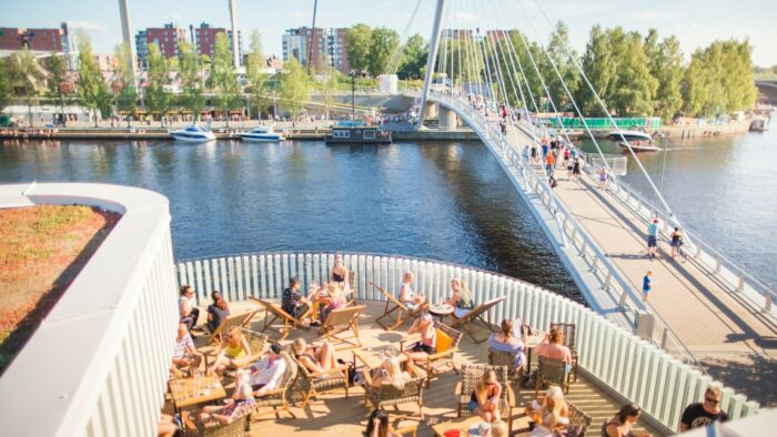 People sit on the patio of a waterside restaurant in front of a bridge that spans the water.