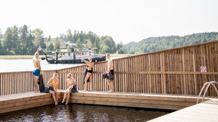 Several people are sitting and standing on a wooden deck that surrounds an outdoor swimming area.