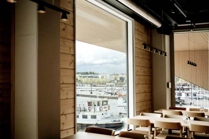 Mesas de restaurante colocadas ao lado de uma janela que vai do chão ao teto com vista para um porto de barcos pequenos.