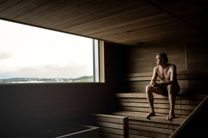 A man sits on a bench in a sauna, gazing out of a long window at the landscape outside.