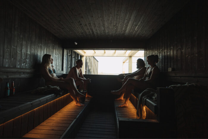 A través de una ventana, la luz penetra en la penumbra de una sauna en la que cuatro personas están sentadas en bancos de madera.