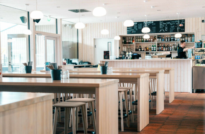 Una hilera de mesas de madera en un restaurante con grandes ventanales, luminoso y diáfano.
