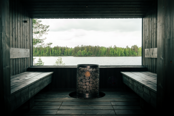 On découvre une vue intérieure d'un sauna vide de tout occupant, avec une large vitre laissant voir un lac et des arbres à l'extérieur.