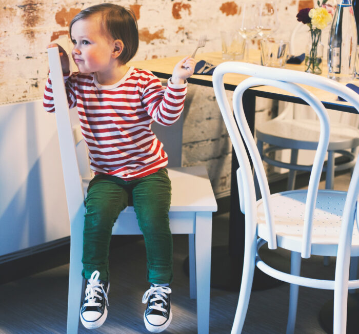طفل يبلغ من العمر أربع سنوات تقريبًا يرتدي ملابس صيفية يجلس على كرسي بجانب طاولة مطعم.