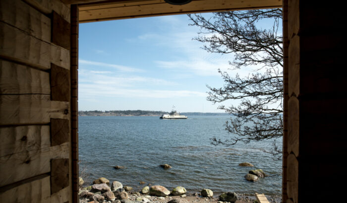 Von der Veranda eines Holzgebäudes aus sehen wir eine weite Wasserfläche mit einem Boot und einer Insel im Hintergrund.