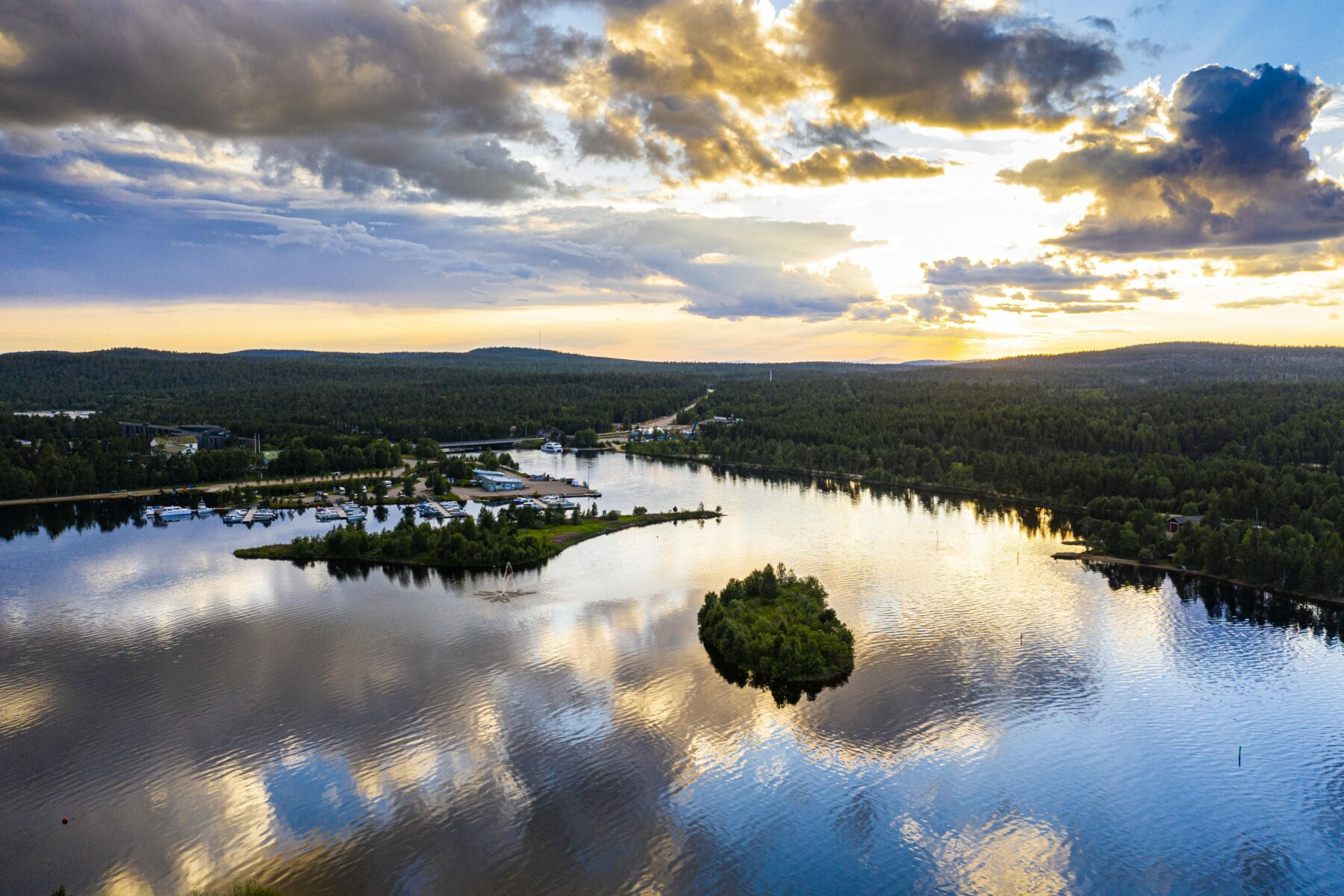 In an aerial shot, the water of a forest-lined lake reflects the sky and clouds.