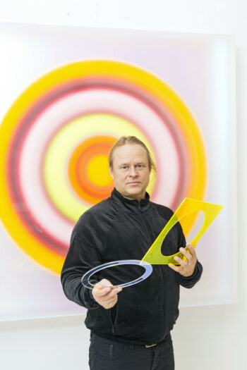 A man holds up flat, ring-shaped plastic forms while standing in front of a wall that displays an artwork formed by a pattern of concentric circles.