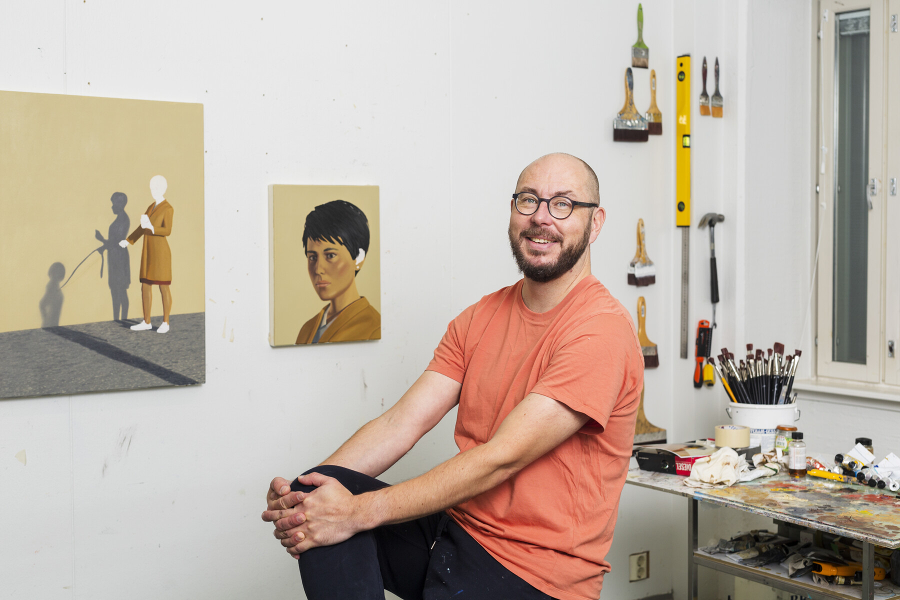 Um homem está sentado em um estúdio de arte com pincéis, ferramentas e pinturas penduradas na parede.