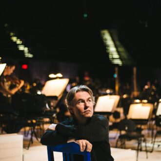 A man dressed in black is looking up thoughtfully, while sitting on a chair in a dimly lit hall with an orchestra setup behind him.