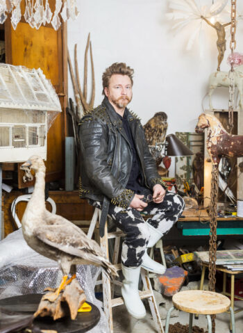 A man in a leather jacket sits on a stool in a cluttered art studio that includes a taxidermically stuffed goose.