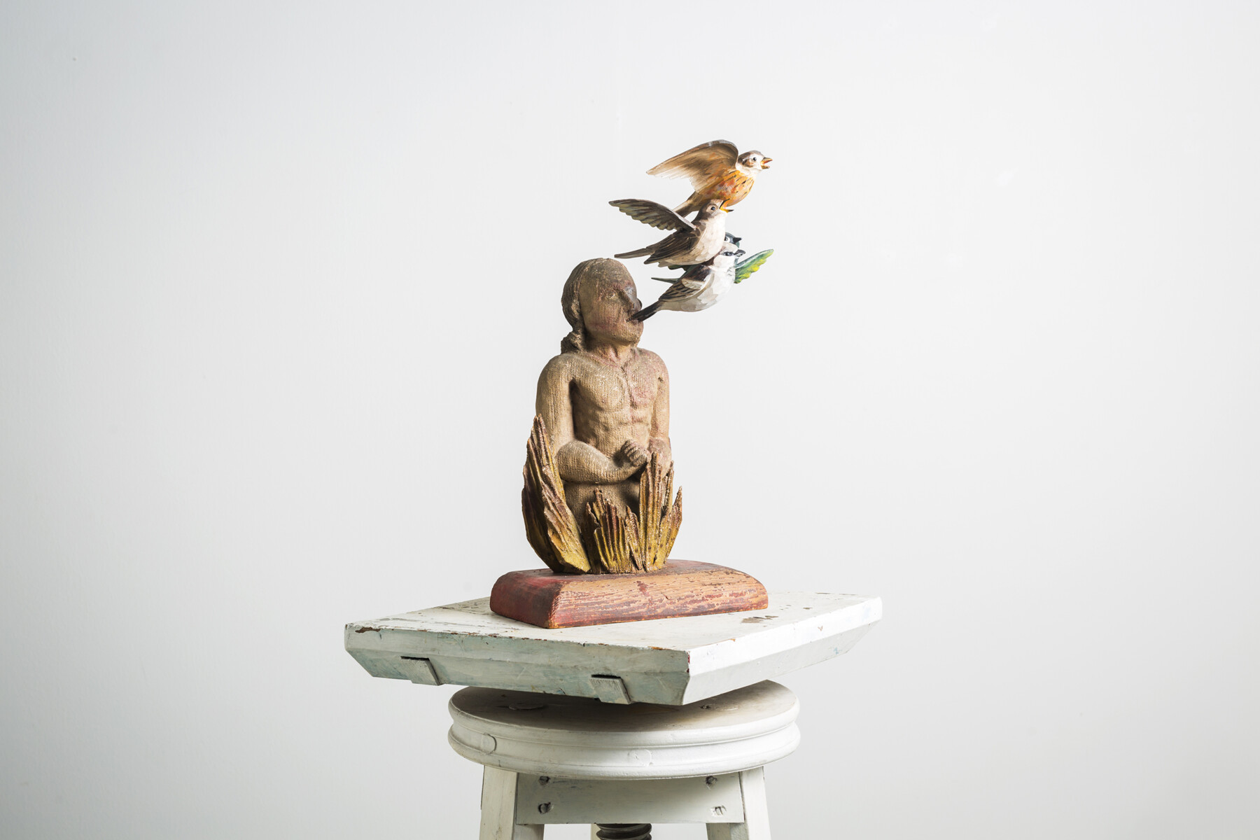 Um pedestal contém a escultura de uma pessoa com vários pássaros coloridos voando perto de sua cabeça ou possivelmente saindo de sua cabeça.