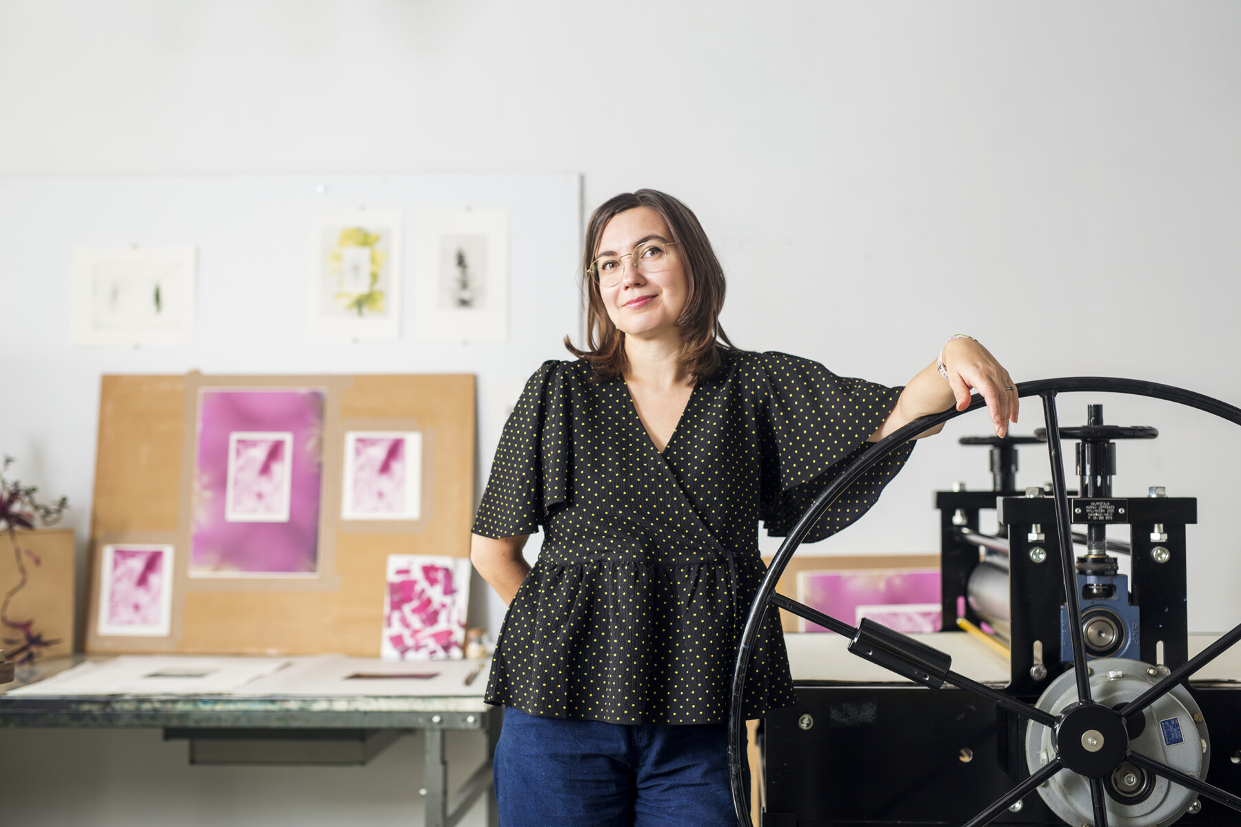 Em um estúdio de arte com gravuras penduradas na parede, uma mulher apoia um braço na manivela de uma impressora antiga.