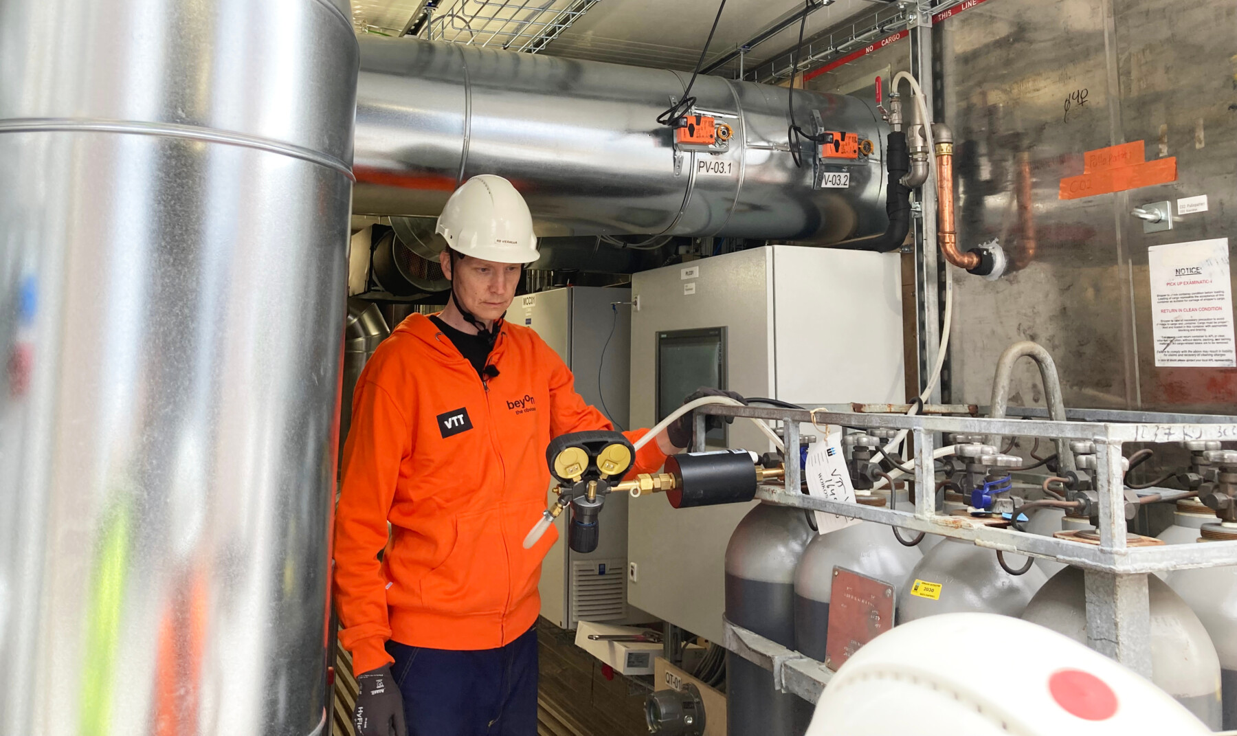 Um homem com capacete e jaqueta colorida observa um conjunto de tubos, tanques e válvulas em uma instalação industrial.