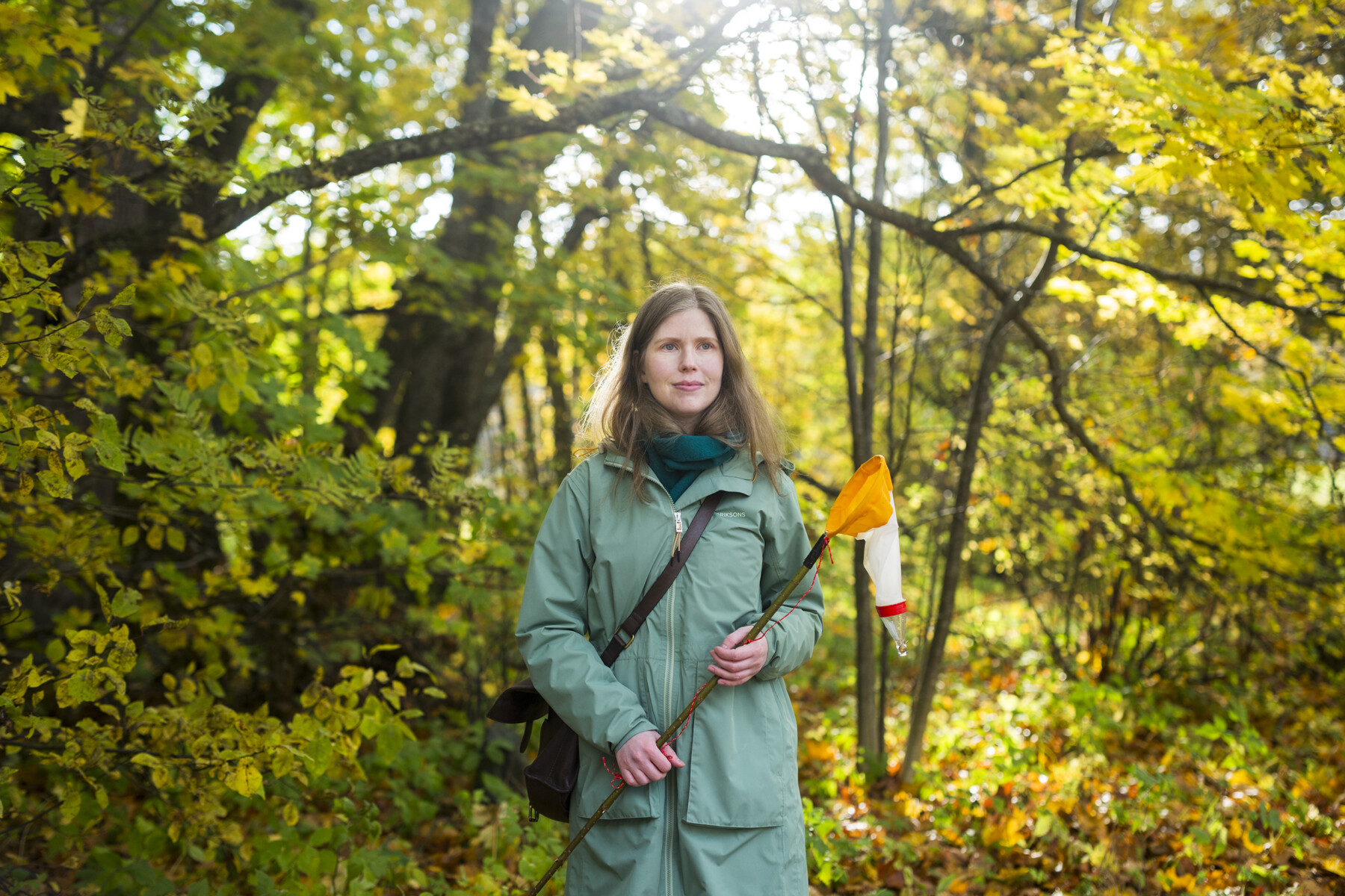 Женщина стоит посреди леса, держа в руке небольшого размера сачок на длинной ручке.