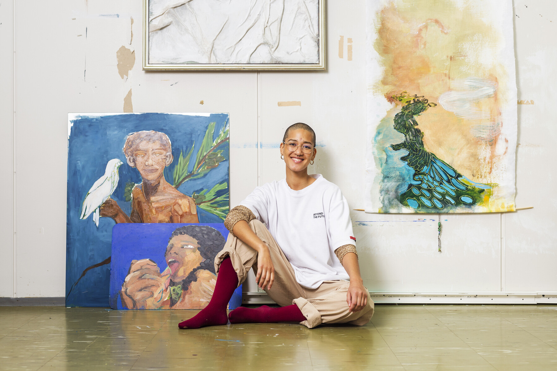 Eine Person mit sehr kurzem Haar sitzt auf dem Boden eines Ateliers mit Bildern an der Wand. 