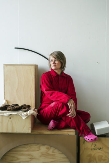 Женщина в красной одежде и розовых тапках-кроксах сидит на деревянном ящике.