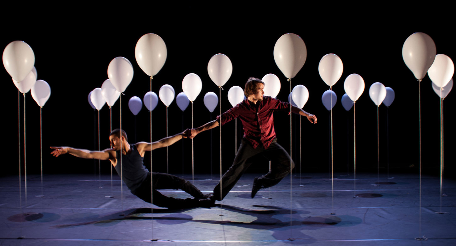 Two men dance among a small forest of white balloons that float above a stage, each with a hanging string that is attached to the floor.