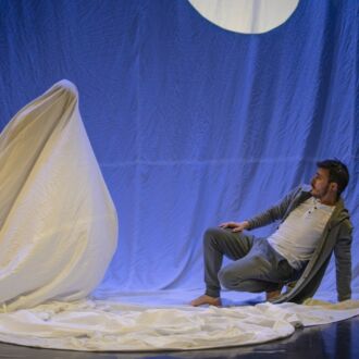 On a stage, a man is squatting and leaning back away from a ghostly figure shrouded in white fabric.