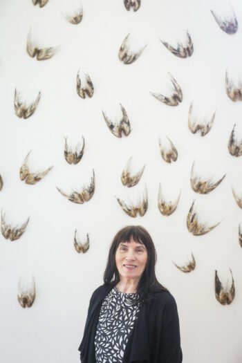 Uma mulher está em frente a uma parede onde estão penduradas dezenas de pares de asas de pássaros.